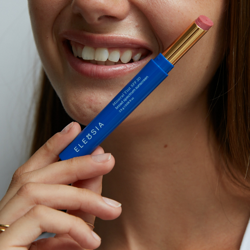 Woman holding a blue makeup product with 'Eleusia' branding near her face.